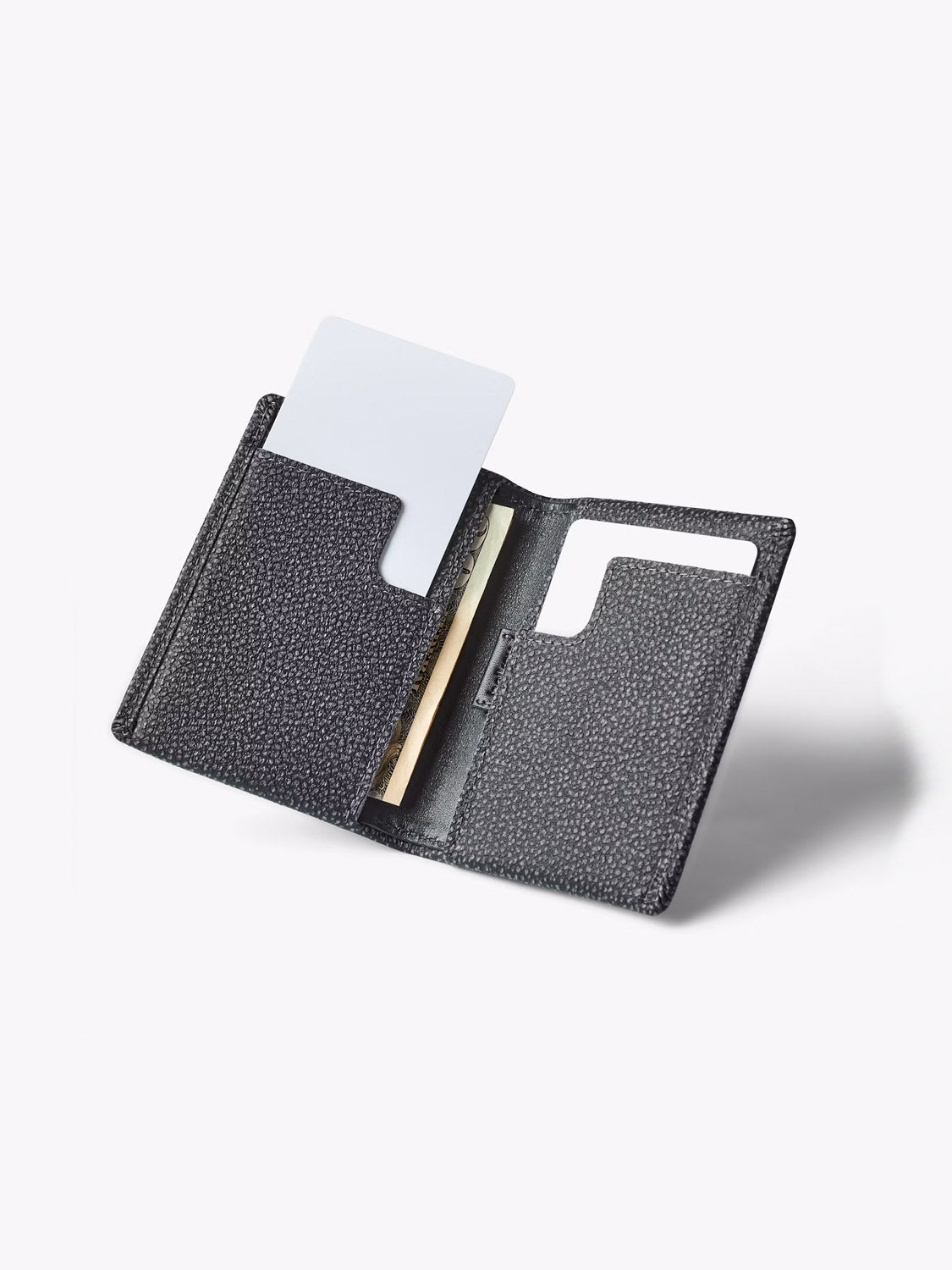The Bellroy Slim Sleeve Wallet in black textured leather is shown open, revealing a white card in one pocket and a few banknotes in another slot, set against a plain light background.