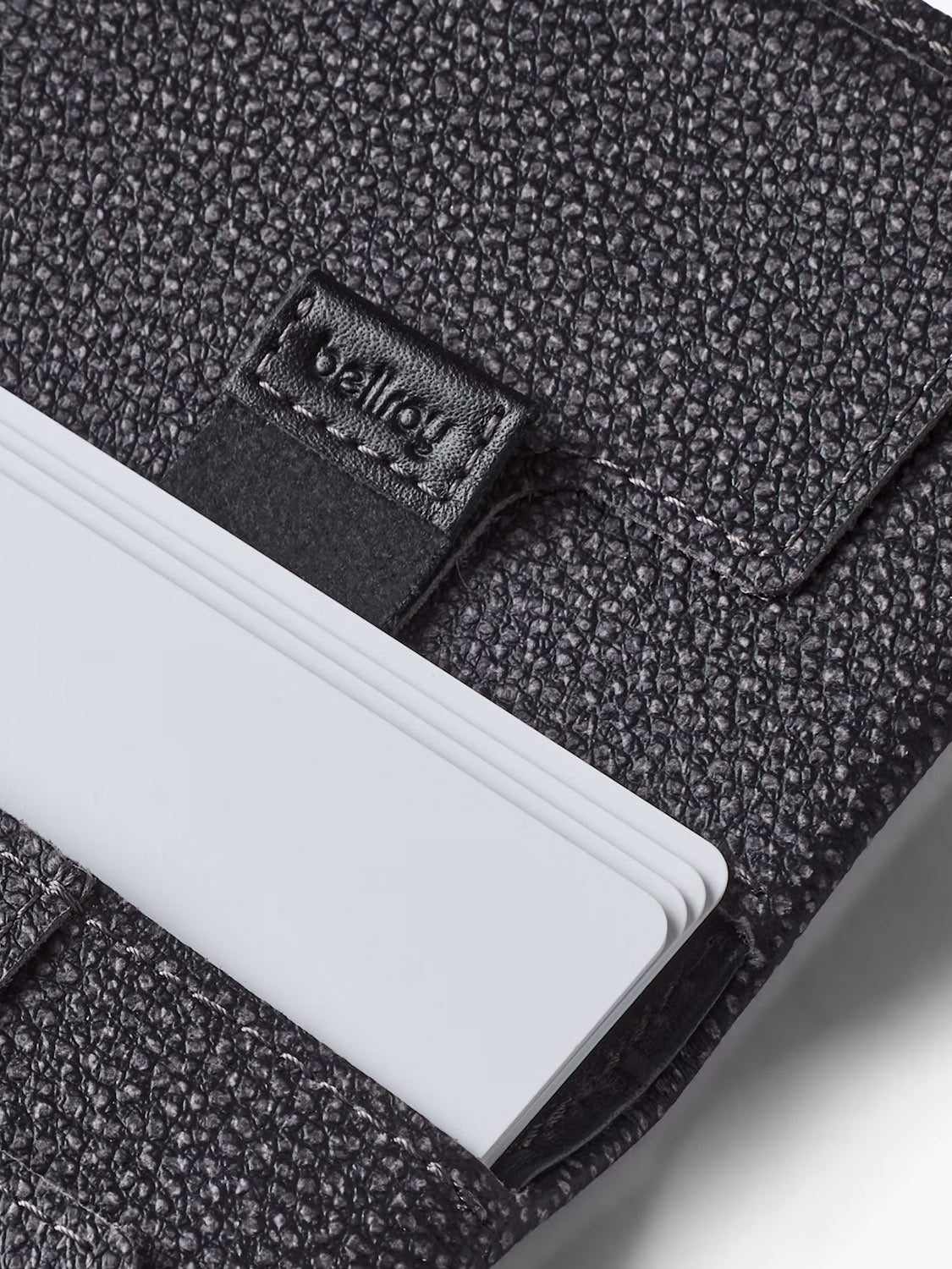 Close-up of Bellroy’s Slim Sleeve Wallet in textured black leather, featuring the brand logo on a flap and several white cards partially inserted in its slot—a truly pocket-friendly accessory.