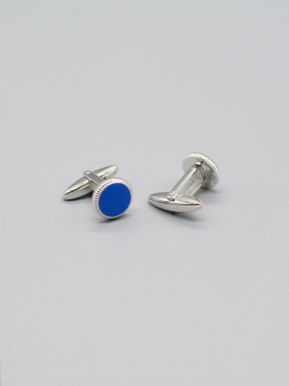 These exquisite Rhodium Plated Steel Enamel Cufflinks by ID Menswear feature round blue centers with colored enamel inlay and a plain gray background. One cufflink is flat while the other stands upright, showcasing their refined elegance.