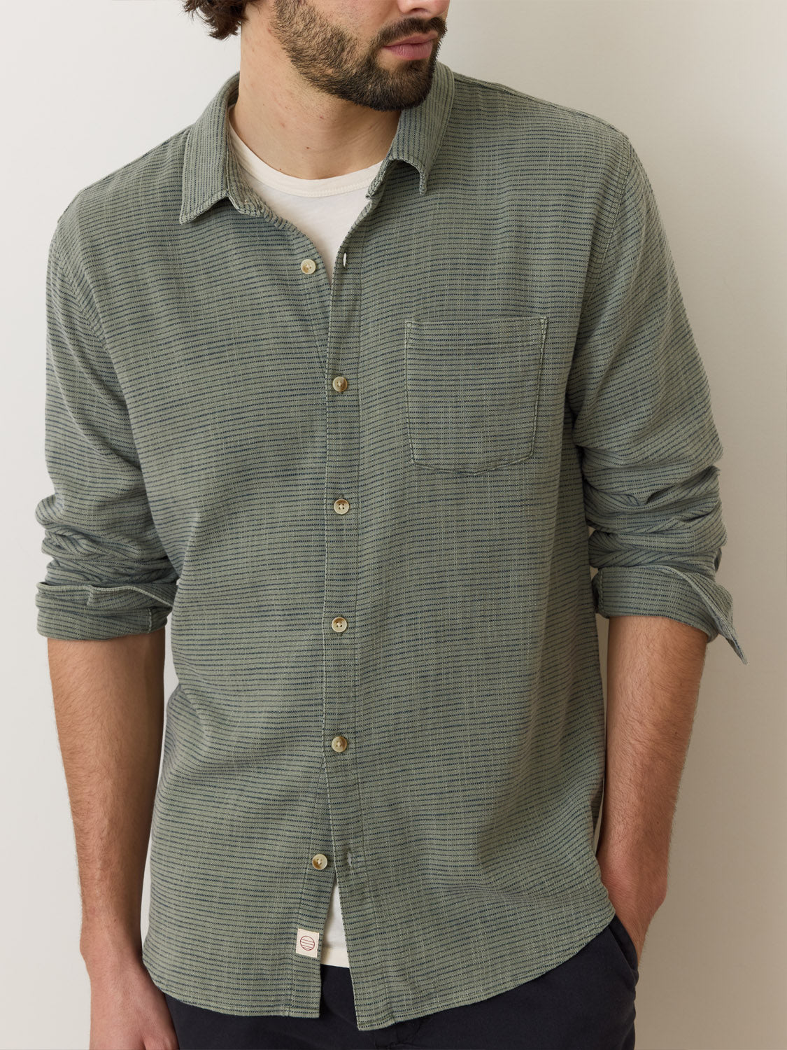 A man in a green Marine Layer Stretch Selvage Long Sleeve Shirt, rolled sleeves revealing added stretch, stands with hands in pockets over a white t-shirt. The shirt complements his beard and short hair as he gazes to the side against a simple backdrop, embodying the relaxed Marine Layer style.