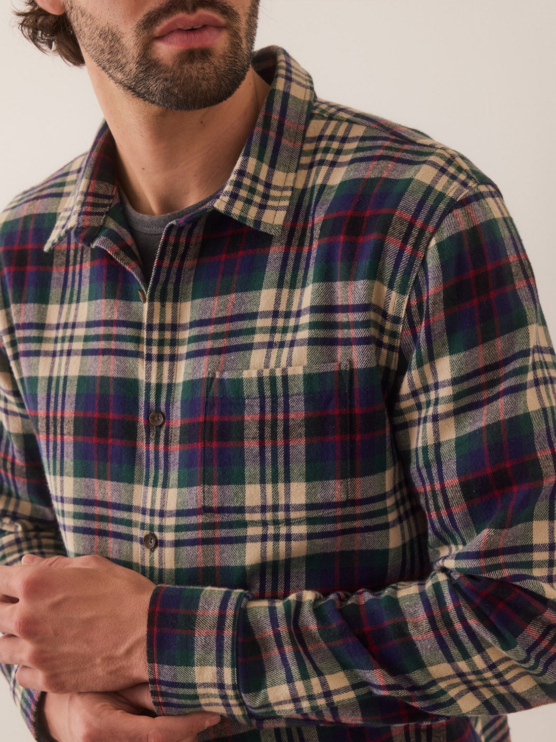 Wearing a Marine Layer Tahoe Flannel Shirt, a person with a beard stands against a plain background. The 100% cotton plaid shirt in green, navy, and beige checks has brushed softness. With arms crossed and face slightly turned away from the camera, their attire subtly shines.
