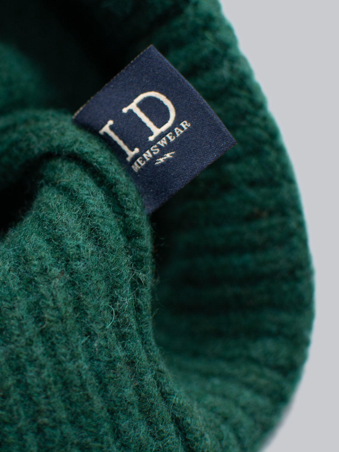 The Telluride Wool Beanie by ID Menswear features a close-up of a cozy, ribbed knit pattern. It is green with a dark blue label displaying white text, offering natural insulation and a comfortable fit.
