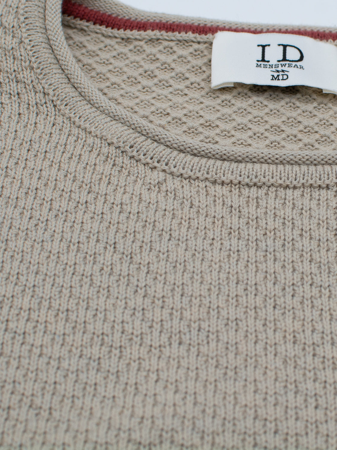 Close-up of a beige textured knit sweater from the Westside Sweater collection, featuring a round neckline. A label inside reads ID Menswear, and a hint of red fabric is visible behind it.