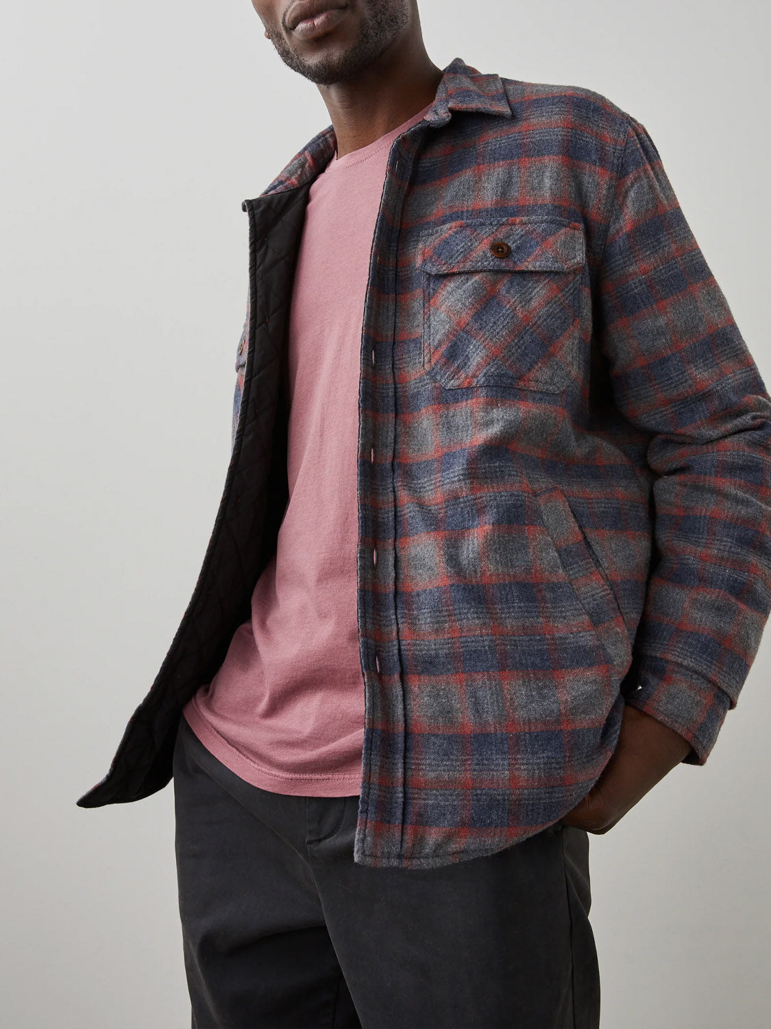 Wearing the Rails Worthing Jacket—a red and gray plaid brushed cotton shirt jacket—over a pink t-shirt and black pants, a person stands with one hand in their pocket against a plain light background.