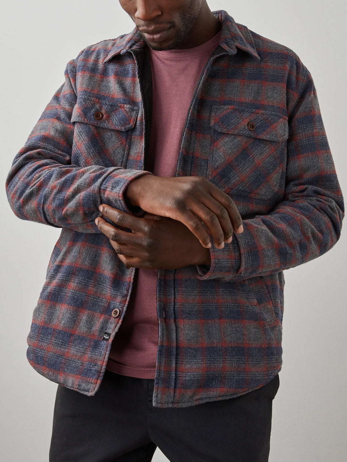 A man wearing the Rails Worthing Jacket—a gray and red plaid brushed cotton shirt jacket—over a mauve sweatshirt adjusts his cuff; only his torso and part of his face are visible against a plain light background.
