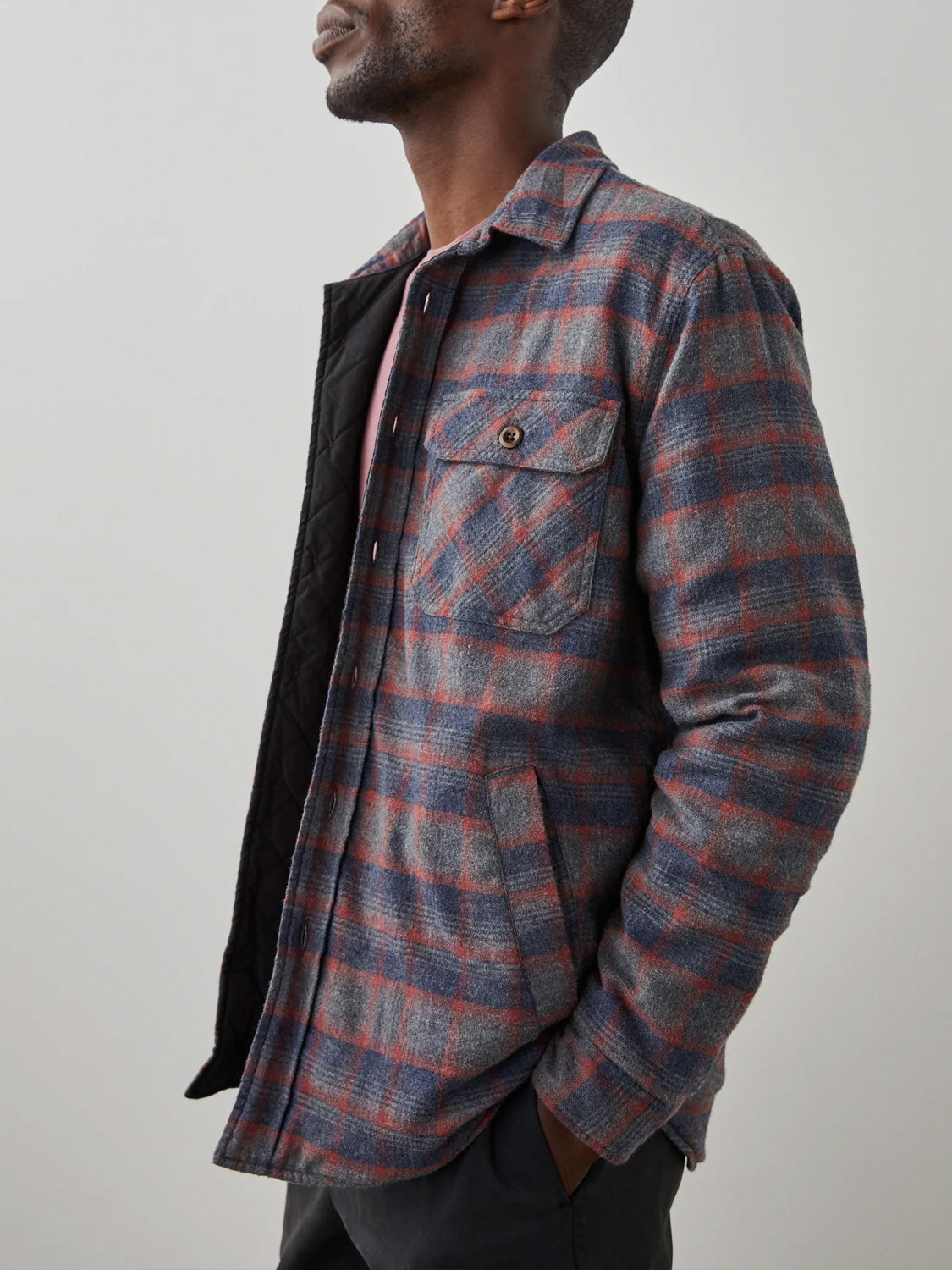 A man stands sideways, looking up, wearing the Rails Worthing Jacket in gray and red plaid brushed cotton with a front pocket. The open jacket reveals a black quilted lining and a glimpse of a light-colored undershirt.