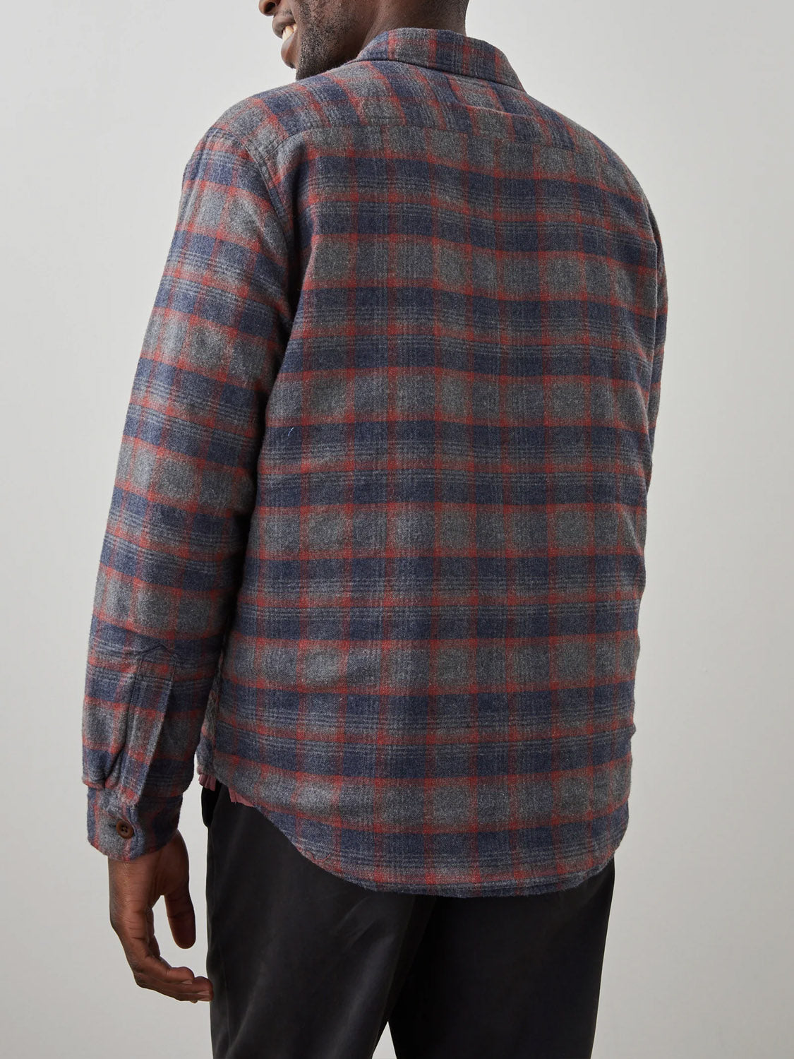 A man is seen from behind wearing the Rails Worthing Jacket, a long-sleeve gray and red plaid shirt jacket made of brushed cotton, paired with black pants. His upper body is shown against a plain light background.