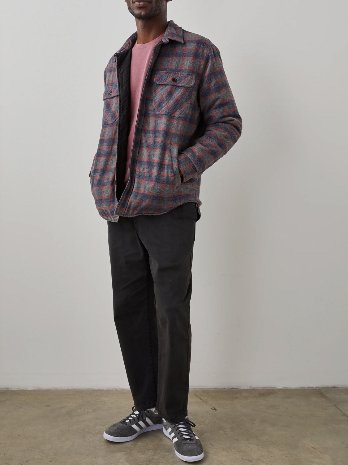 Indoors, a person wears the Rails Worthing Jacket—a gray and red plaid shirt jacket with brushed cotton and quilted lining—over a pink shirt, black pants, and gray Adidas sneakers. Their hands are in the jacket pockets.