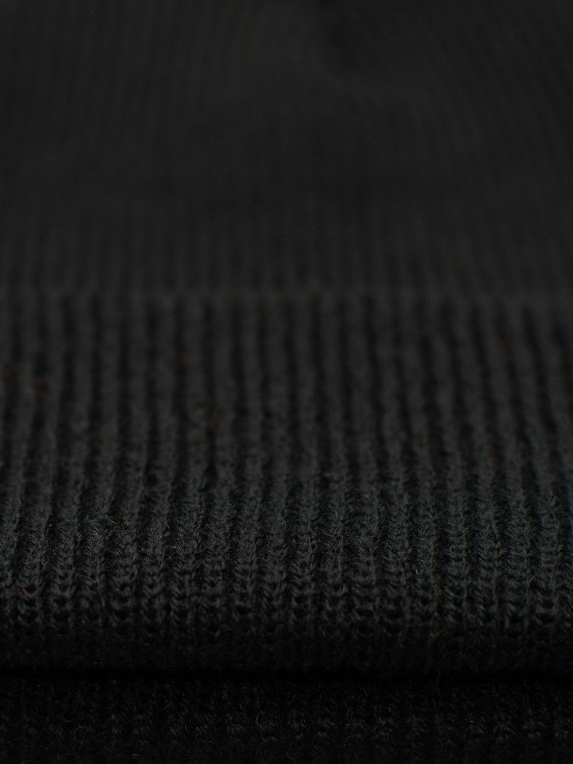 Close-up of the Yonker Ribbed Acrylic Beanie by ID Menswear in dark gray knit fabric. The ribbed texture features horizontal lines, perfect for cozy winter wear. Lighting enhances the weave and structure beautifully.