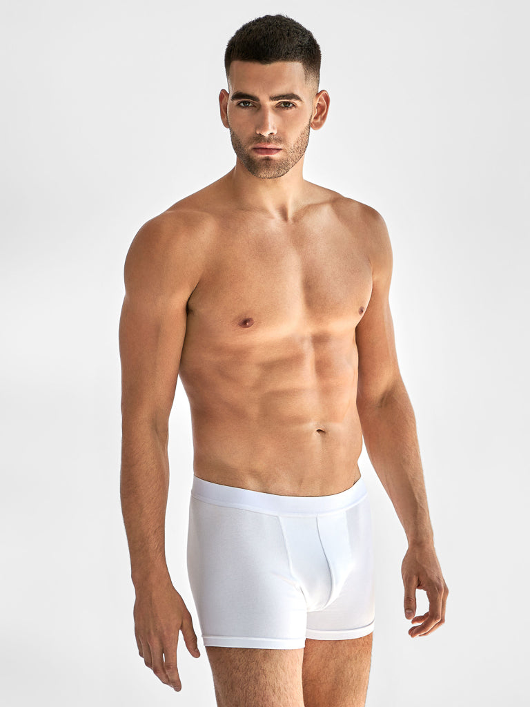 A man with short dark hair poses against a plain backdrop, wearing Bread & Boxers white boxer briefs made from organic cotton. These briefs have an elastic waistband and reinforced front as he stands slightly turned to the side.