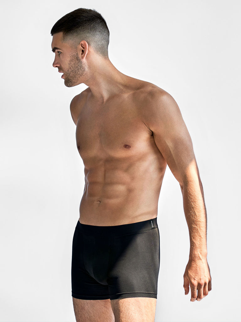 A shirtless man with short hair stands in profile facing left, wearing Bread & Boxers black boxer briefs. Made from soft organic cotton, these briefs feature a sleek design and an elastic waistband for comfort. The background is plain white.