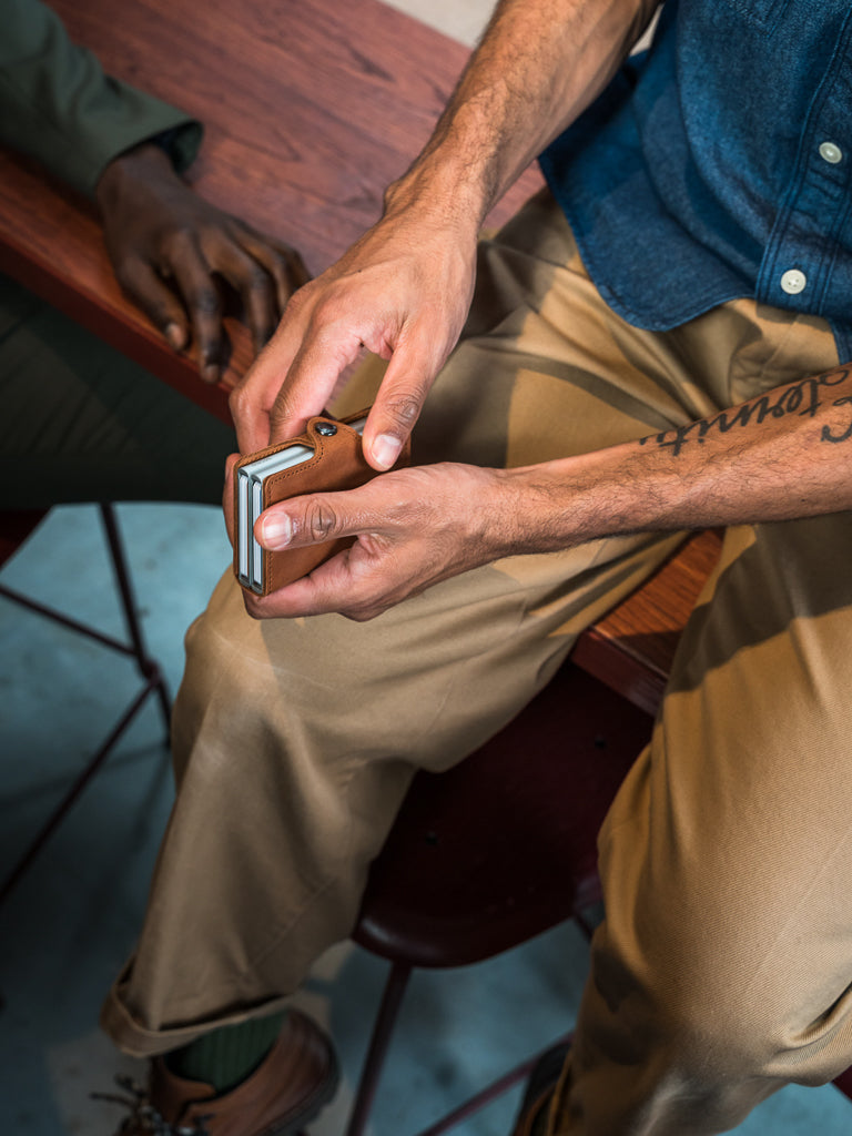 A person in tan pants and a blue shirt shuffles playing cards while sitting on a stool, with a Secrid Twin Wallet Matte Finished Leather and another person's arm visible on a wooden table nearby.