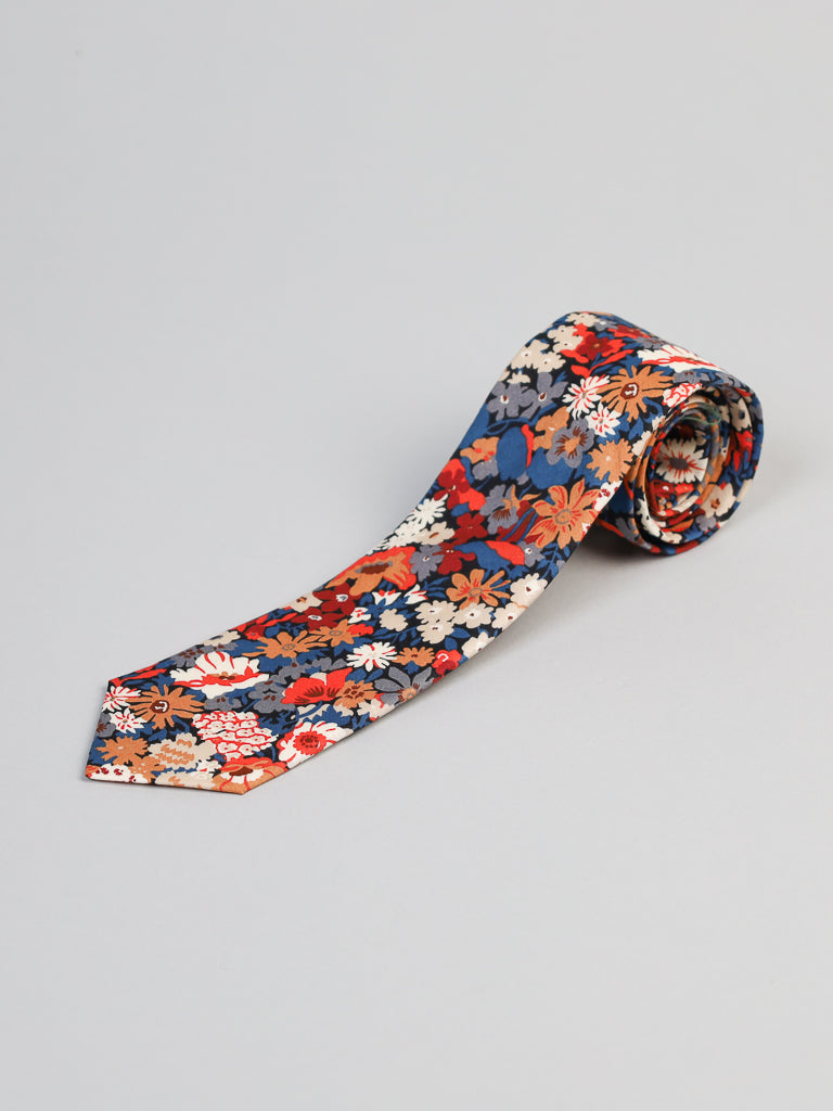 The ID Menswear Liberty of London Tana Lawn™ Kanazawa Tie, featuring a vibrant floral pattern in red, orange, blue, white, and gray Tana Lawn cotton, is rolled up and displayed on a plain light gray background.