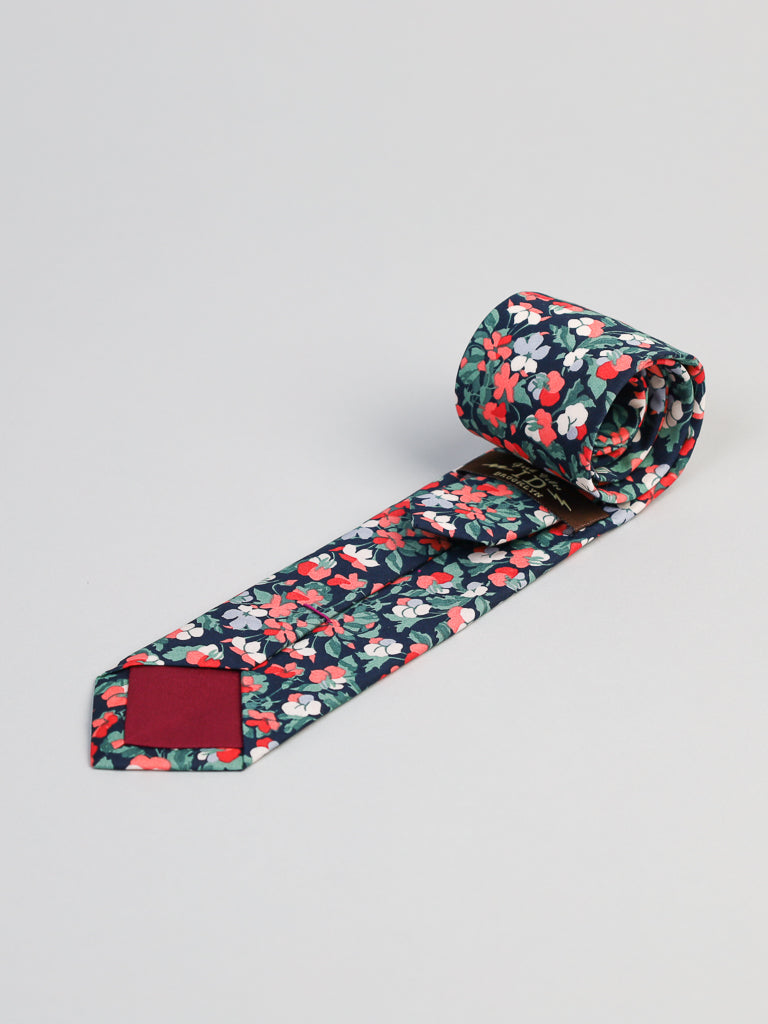 The Liberty of London Tana Lawn™ Classic Tie by ID Menswear features a vivid red, white, and green floral print on dark fabric, handmade in Brooklyn from premium cotton and shown rolled up on a light gray surface.