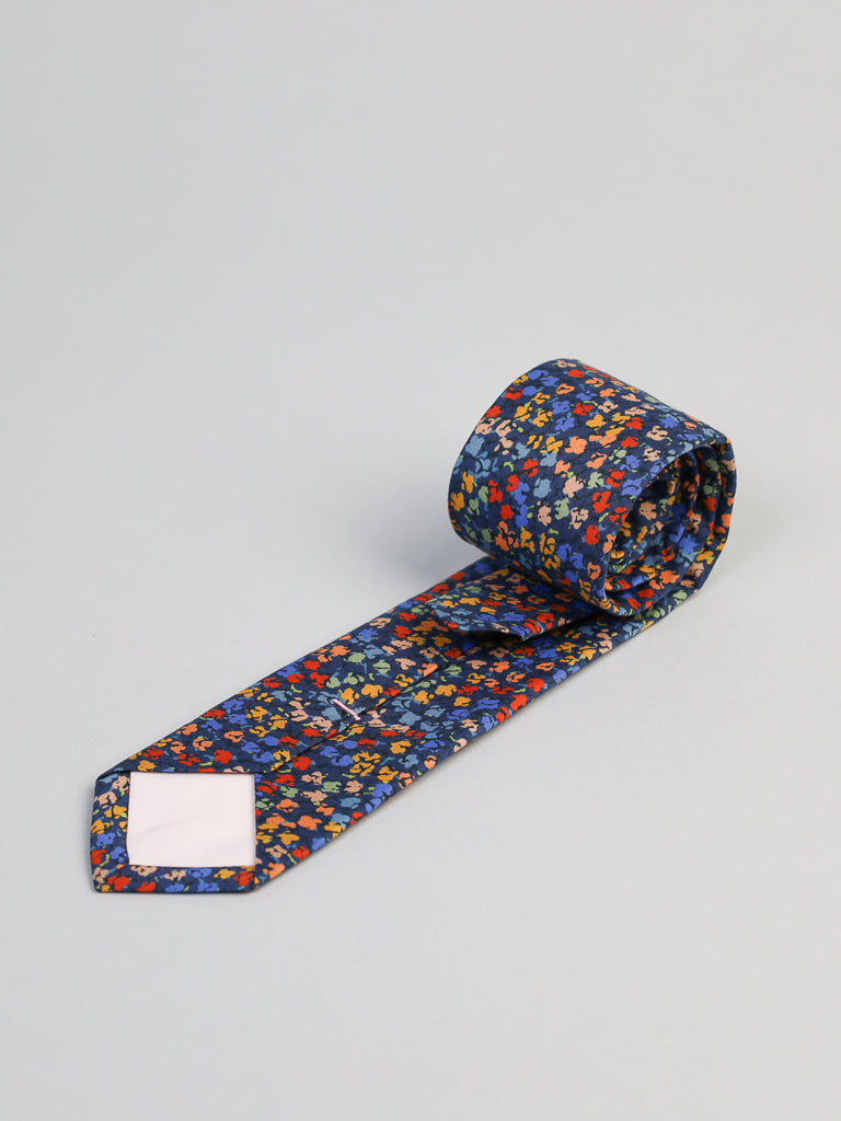The ID Menswear Liberty of London Tana Lawn™ Gizmo Tie, featuring a dark blue base with a red, yellow, and blue floral pattern, is shown rolled up on a plain light gray background.
