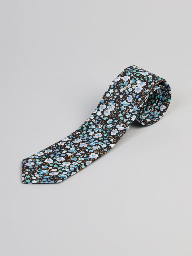 A rolled-up ID Menswear Liberty of London Tana Lawn™ Jo Tie with a blue, white, and light green floral pattern on a dark background, displayed on a plain light gray surface.