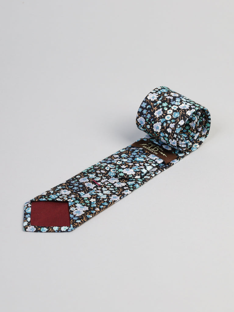 The ID Menswear Liberty of London Tana Lawn™ Jo Tie features a handmade blue, white, and brown floral pattern, crafted in Brooklyn from luxurious cotton and shown rolled up on a light gray background.