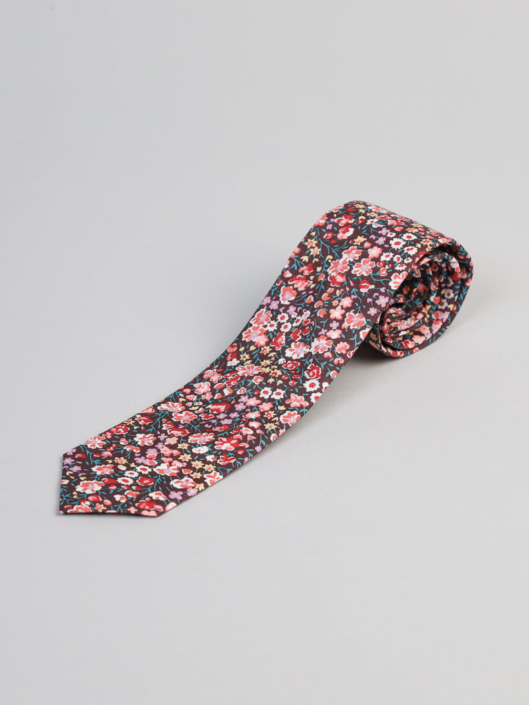 The ID Menswear Liberty of London Tana Lawn™ Jo Tie, handmade in Brooklyn, features a colorful floral pattern in red, pink, and green on a dark background and is displayed rolled up on a light gray surface.