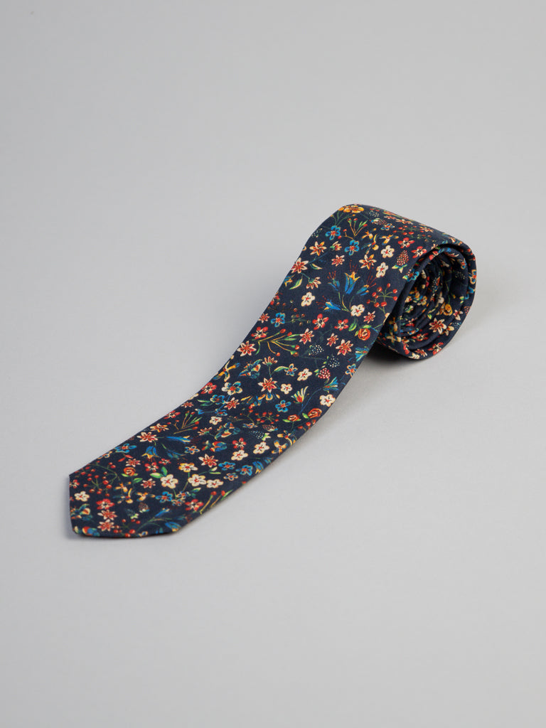 A Liberty of London Tana Lawn™ Donna Tie by ID Menswear, handmade in Brooklyn with a vibrant floral print in red, yellow, blue, and green on cotton, is neatly rolled and displayed on a light gray surface against a dark background.