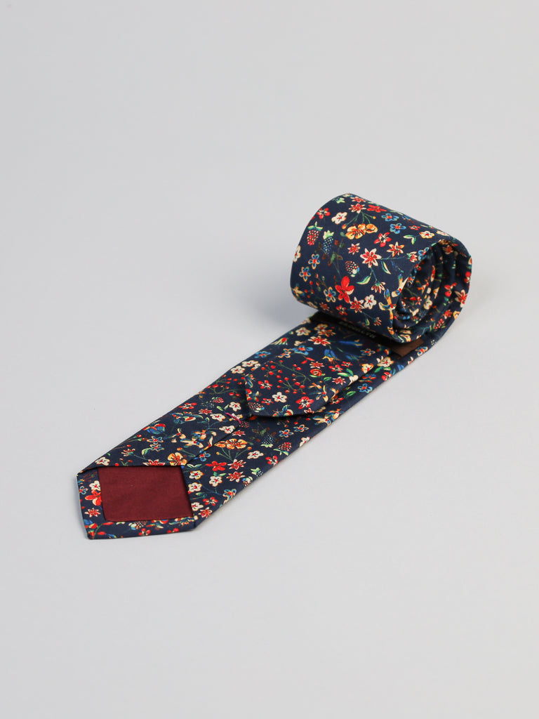 The Liberty of London Tana Lawn™ Donna Tie by ID Menswear, handmade in Brooklyn from luxurious navy floral cotton, features a maroon tip patch and is shown rolled up on a light gray surface.