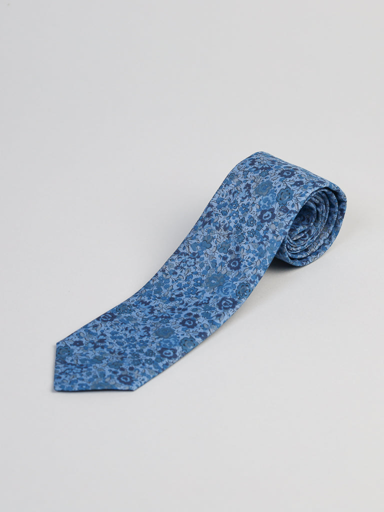 The ID Menswear Liberty of London Tana Lawn™ Georgina Tie, featuring a blue floral pattern, is handmade in Brooklyn and shown neatly rolled on a plain light gray surface.