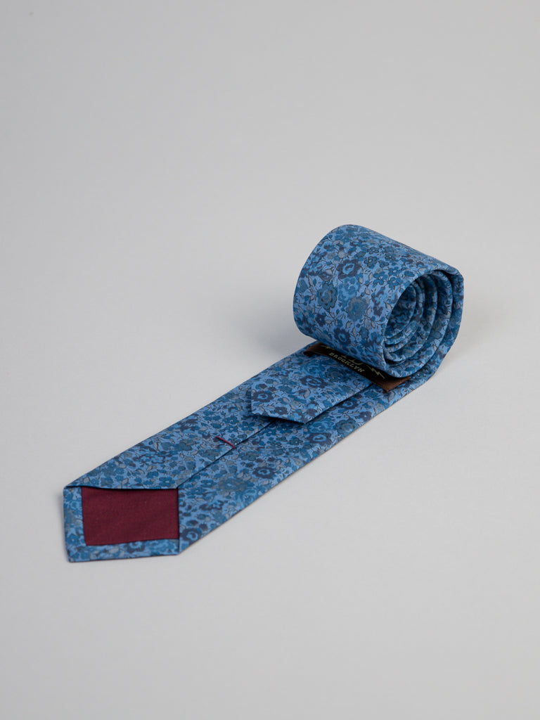 The ID Menswear Liberty of London Tana Lawn™ Georgina Tie, handmade in Brooklyn, features a blue floral pattern and maroon lining at the tip. It’s neatly rolled on a light gray surface and offers a silky touch.