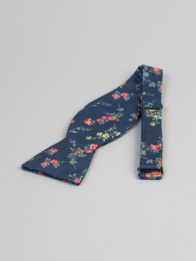 A blue bow tie with a red and yellow floral pattern, crafted from Tana Lawn™ cotton by ID Menswear, rests on a gray surface. The Liberty of London-inspired strap is partially visible beside it.