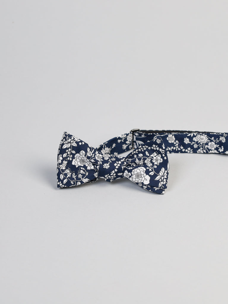 A handmade navy bow tie featuring a white floral pattern, reminiscent of Liberty of London Tana Lawn designs, is styled elegantly against a light gray backdrop. This ID Liberty of London Tana Lawn™ Navy Lawn Cotton Bowties from ID Menswear combines intricate flowers and leaves for a stylish look.