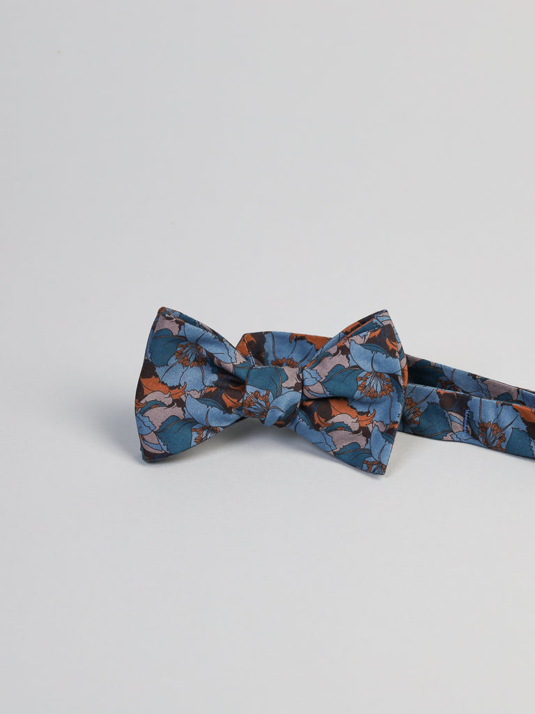 The ID Liberty of London Tana Lawn™ Eustacia Blue Cotton Bowtie by ID Menswear features a floral pattern in orange and dark blue on a light gray surface. This handmade Brooklyn accessory combines style and craftsmanship with its elegant, silk-like touch.