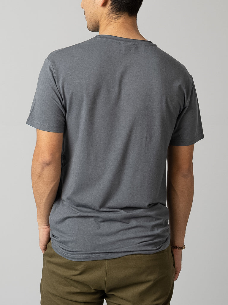 A person in an ID Menswear Bello Bamboo Blend Rolled Neck Tee and olive green pants is facing away from the camera. Hands in pockets, they exude a cool vibe against the plain gray background.