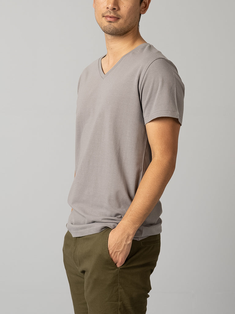 A man embodies a casual look in a Form V Neck Tee by ID Menswear, light gray and form-fitting, paired with olive green pants. He stands with hands in his pockets against a plain gray background, head slightly turned downward.
