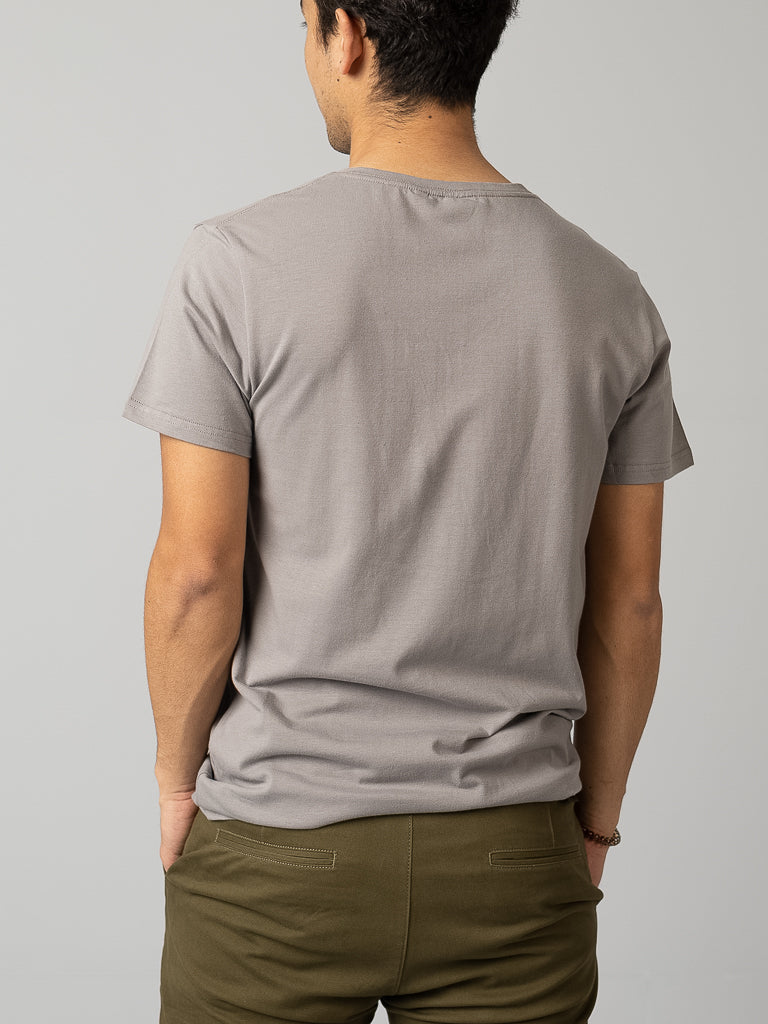 A person with short dark hair stands in a form-fitting gray Form V Neck Tee by ID Menswear, paired with olive green pants. Against a light gray background, hands in pockets, they epitomize effortless style for any casual occasion.