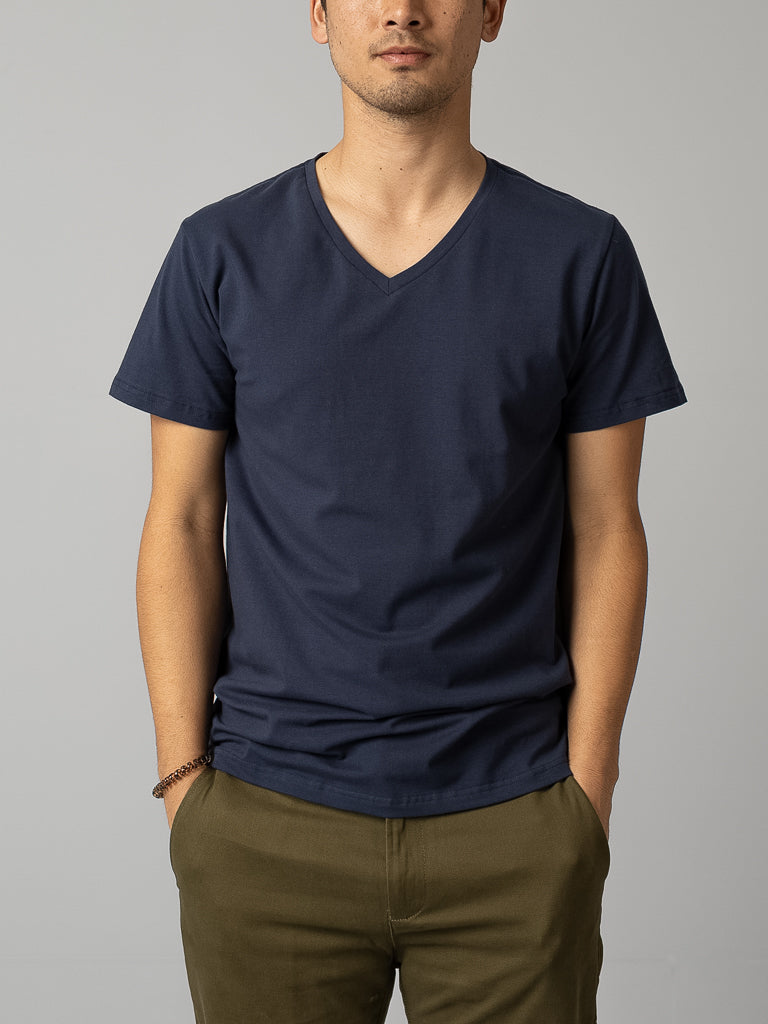 A person in a form-fitting Form V Neck Tee by ID Menswear in navy blue and olive green pants stands with hands in pockets against a light gray background, ideal for casual occasions. Only the upper body is visible.