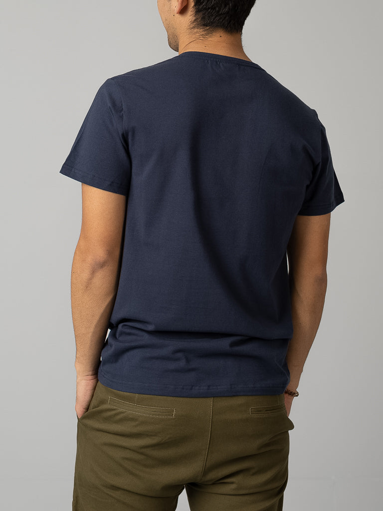 A person stands with their back to the camera, wearing a navy Form V Neck Tee by ID Menswear and olive green pants, projecting a casual vibe. Their left hand is in their pocket against a neutral background.