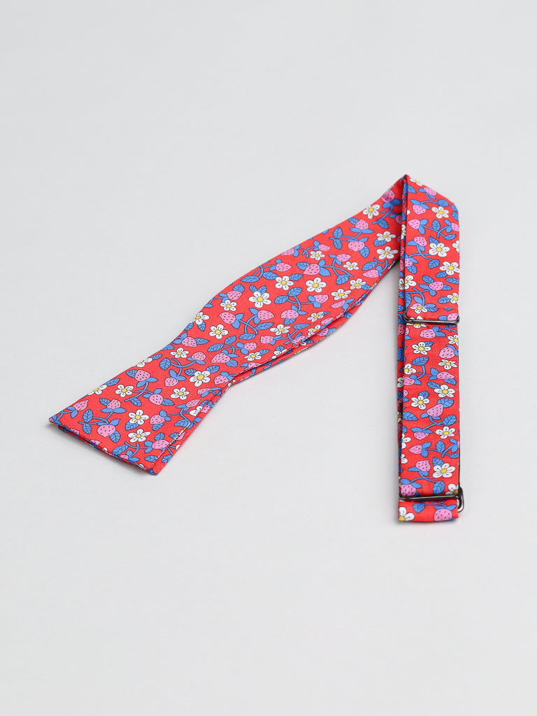 The ID Menswear bow tie, made from Liberty of London Tana Lawn™ cotton with a floral pattern, features blue and white flowers on red. Handmade in Brooklyn, it elegantly rests on a light gray surface.