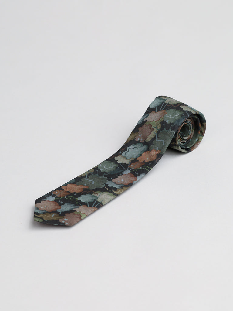 A rolled-up Liberty of London Tana Lawn™ Cloud Nine Tie by ID Menswear, handmade in Brooklyn from cotton, displays a green, brown, and gray leaf pattern on a dark background atop a plain white surface.