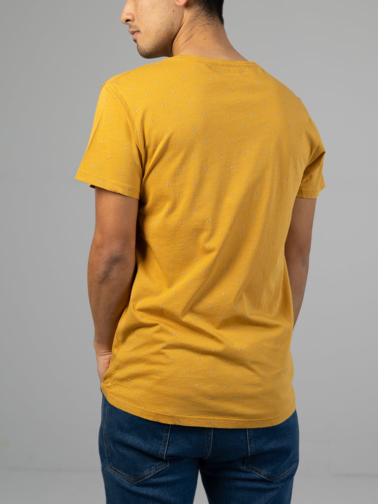 A person facing away dons the Heritage Printed Organic Cotton Tee by ID Menswear, featuring a classic fit in mustard yellow with subtle dotted patterns, paired with blue jeans against a plain light gray backdrop.