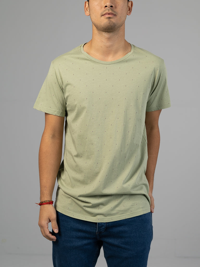 A person in an ID Menswear Heritage Printed Organic Cotton Tee, featuring a light green hand screen printed classic fit with small dots, and blue jeans stands against a gray background. A red bracelet adorns one wrist, and their face is not fully visible in the photo.
