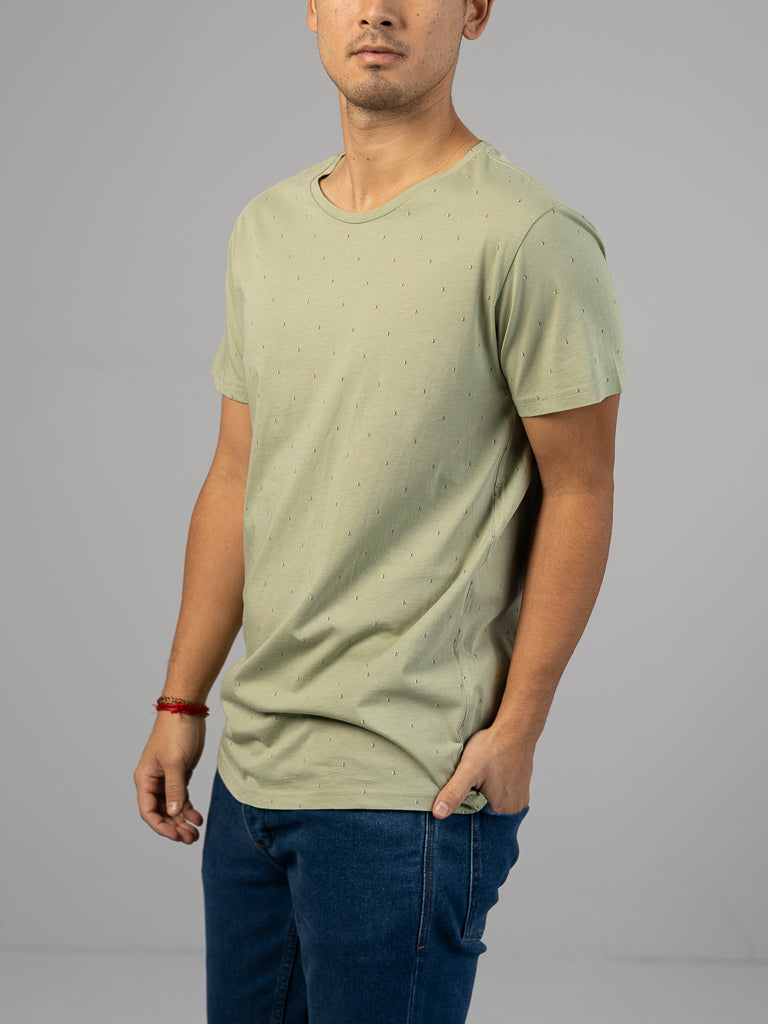 A person in ID Menswear’s Heritage Printed Organic Cotton Tee, featuring a light green hand screen-printed design with small dots, pairs it with blue jeans against a gray backdrop. The classic fit T-shirt is short-sleeved, and they have their right hand in their pocket.