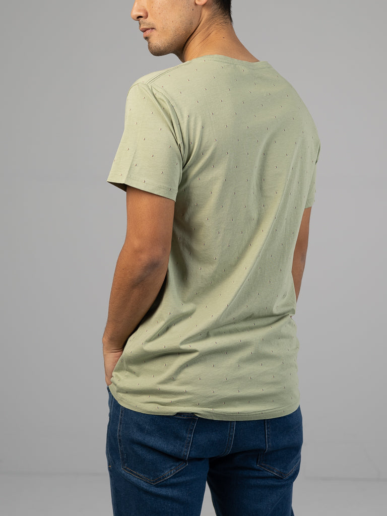A person stands with their back turned, wearing an ID Menswear Heritage Printed Organic Cotton Tee in light green and classic-fit dark blue jeans against a plain gray background.