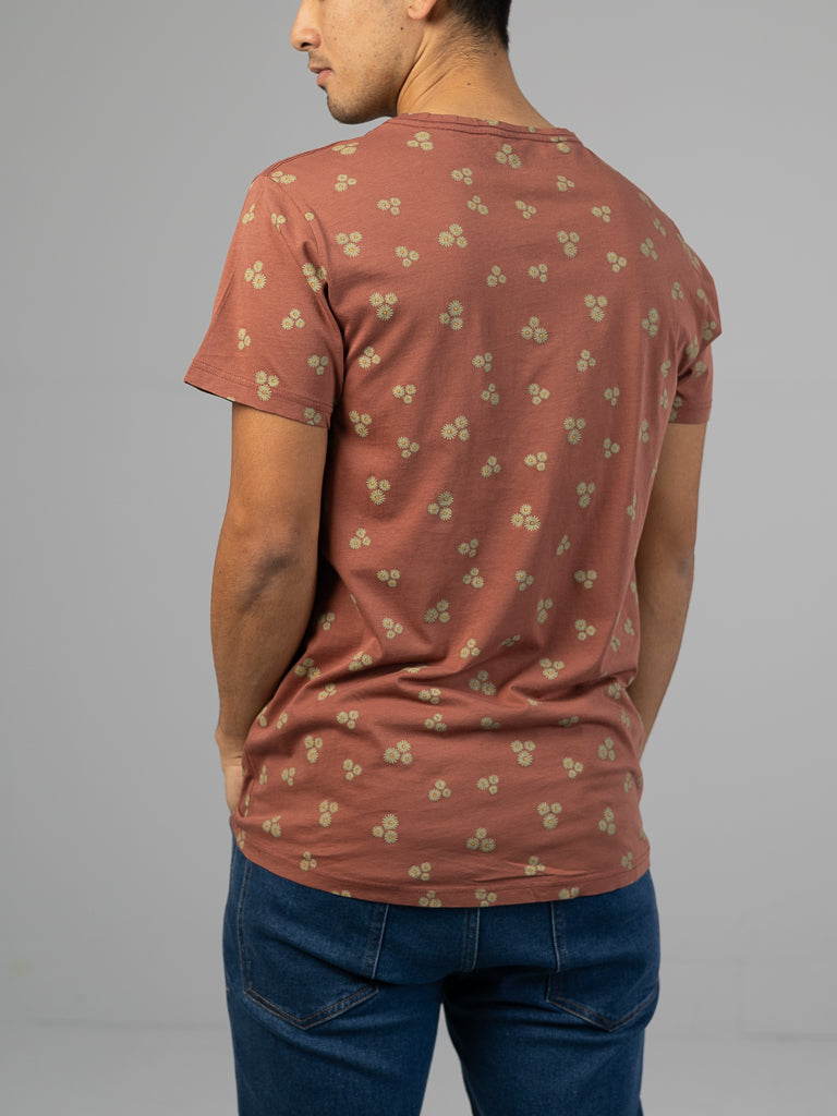 A person wearing the Heritage Printed Organic Cotton Tee by ID Menswear, featuring a classic fit and subtle floral print on organic cotton in brown, alongside blue jeans, stands with their back turned against a plain, light background that highlights the hand screen printed design.