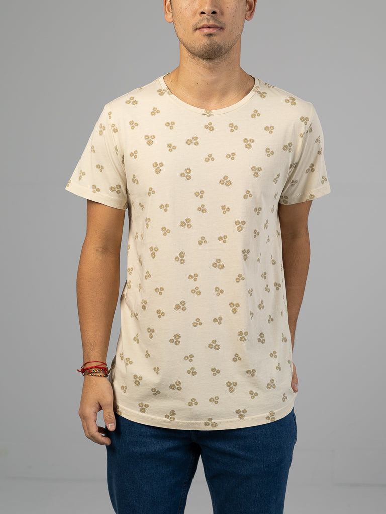 A person stands against a plain gray background wearing ID Menswears Heritage Printed Organic Cotton Tee, featuring a classic fit, cream color, small repeating floral pattern, hand screen printing, round neck, and short sleeves paired with blue jeans. They have a red bracelet on their wrist.