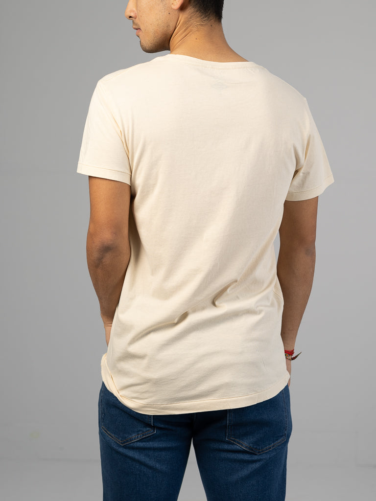 An individual is seen from behind, dressed in a plain cream Legacy Organic Cotton Tee by ID Menswear and blue jeans, their hands in pockets. The classic fit harmonizes with the understated gray background beautifully.