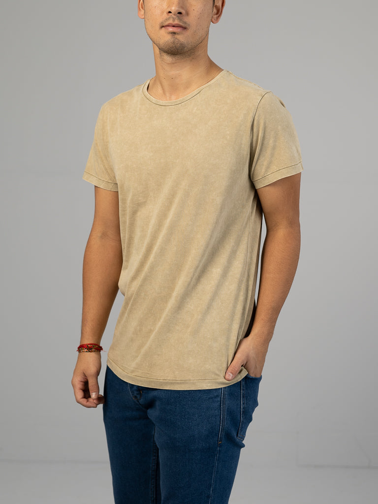 Against a plain gray backdrop, a person in ID Menswears Legacy Organic Cotton Tee in light beige—complementing their vintage style with blue jeans—stands casually with one hand in their pocket. Their style is accentuated by a red beaded bracelet on their wrist.