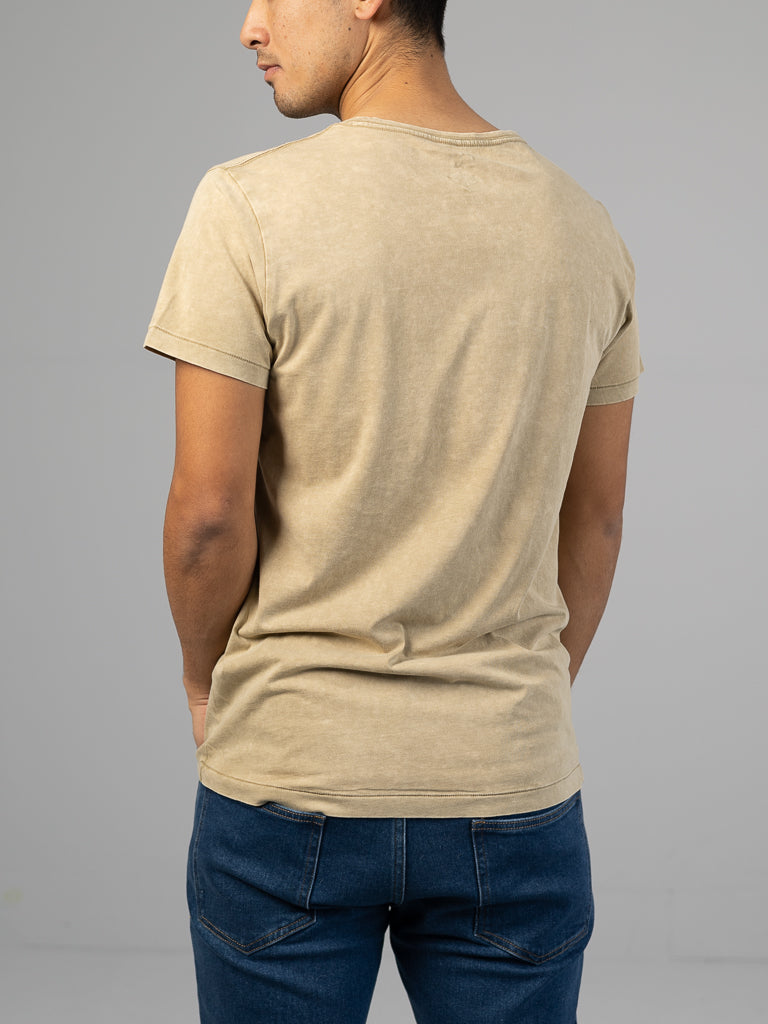 Facing away from the camera, a person wears an ID Menswear Legacy Organic Cotton Tee in faded beige with a vintage look, paired with dark blue jeans. The tee boasts a classic fit against a plain, light gray background.
