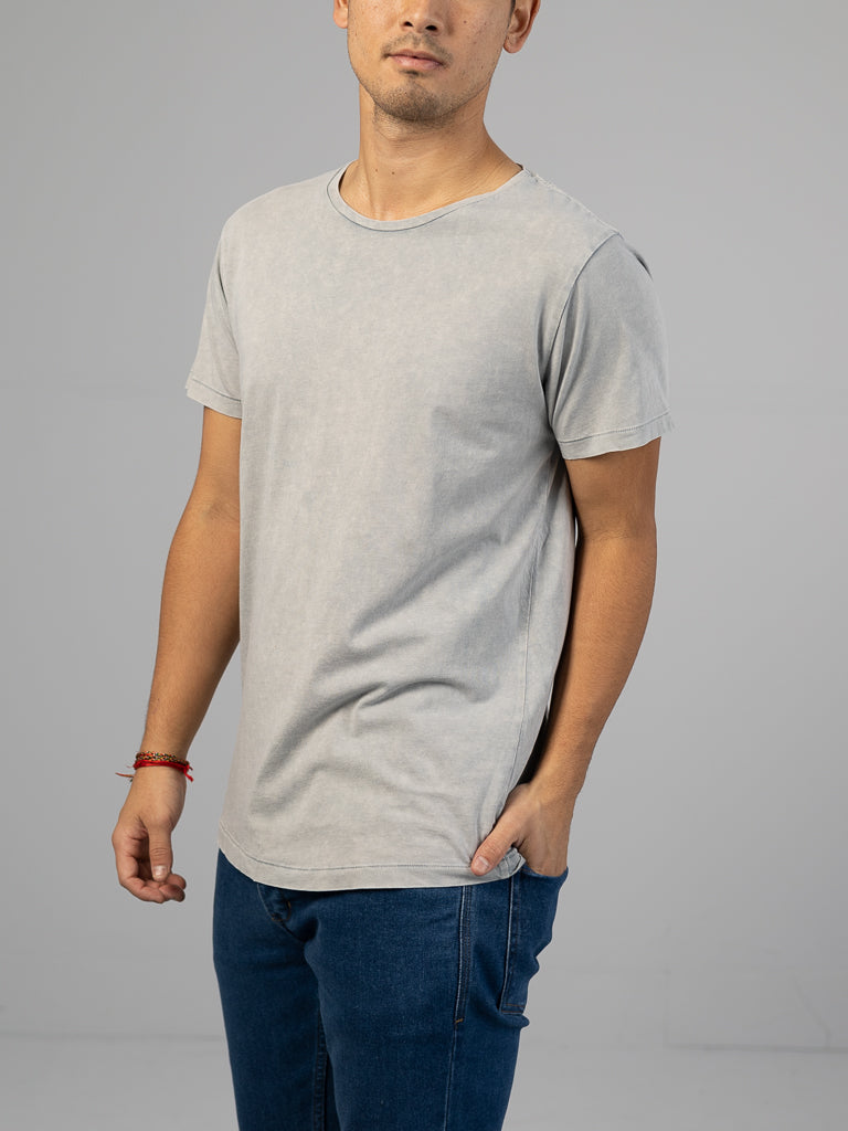 Wearing ID Menswears Legacy Organic Cotton Tee in light gray with blue jeans, a person stands against a plain gray background. Their left hand is in the pocket, and a red bracelet decorates their right wrist. The relaxed vibe is enhanced by the organic cotton tee, though their face is mostly cropped out.
