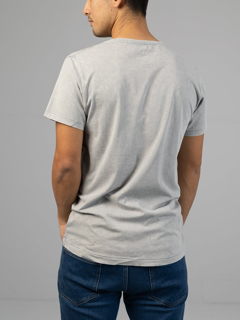 A person in an ID Menswear Legacy Organic Cotton Tee and dark blue jeans faces away, set against a plain gray background that enhances the vintage vibe of their outfit.