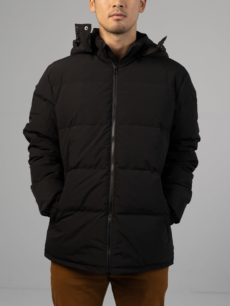 A person wearing the ID Menswear Arctic Black Puffer Down Jacket with a hood stands with hands in pockets against a plain light gray background. The individual's face is partially visible.