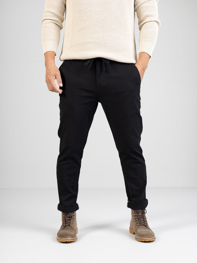 A person in a cream sweater and Kent Drawstring Canvas Pant by ID Menswear stands against a light grey background, wearing tan lace-up boots with hands casually in pockets. The image is cropped from the shoulders up, highlighting a sleek, contemporary style.