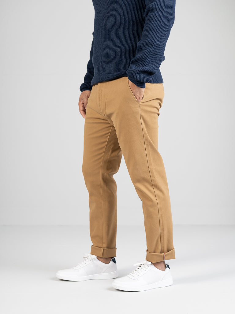 A person in a navy sweater, ID Menswears regular slim-fit Banker Twill Stretch Chinos with rolled cuffs, and white sneakers stands against a plain light background, hands in the chinos mid-rise pockets.